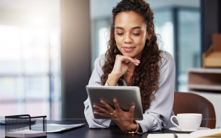 Woman smiling while using a tablet at a desk with coffee and notebook.