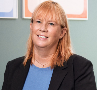 Märit Sareyko, CEO of Epical, smiling at the camera in an office setting with a green wall and colorful posters in the background. She is wearing a blue top and a black blazer, standing next to a green plant.