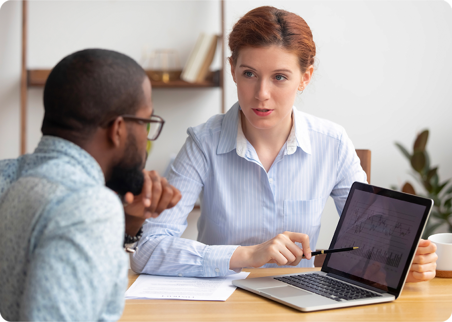 Two colleagues sitting at a desk, with one pointing at a laptop screen displaying charts and graphs during a discussion.