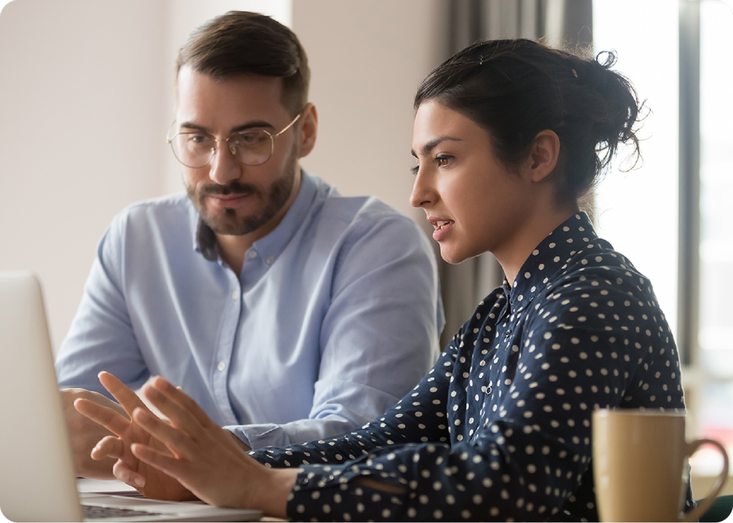 Two colleagues discussing work while looking at a laptop, with a coffee mug on the desk.