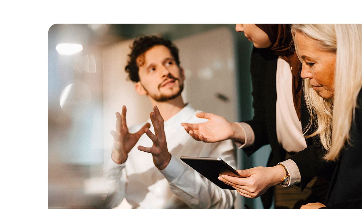 Three colleagues in a meeting, one speaking with hand gestures while the others look at a tablet and listen attentively.