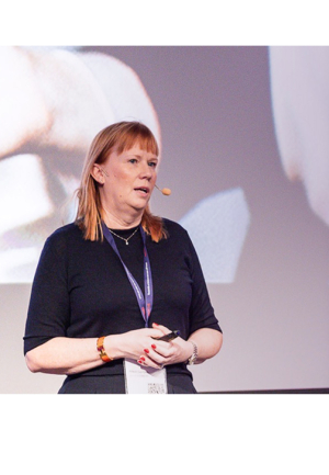 Marit Sareyko, wearing a black top and skirt, speaks on stage with a headset microphone. She is presenting at a conference in front of a large projected image. She has a lanyard with a name badge and is holding a small device in her hands.