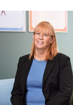 Märit Sareyko, CEO of Epical, smiling at the camera in an office setting with a green wall and colorful posters in the background. She is wearing a blue top and a black blazer, standing next to a green plant.