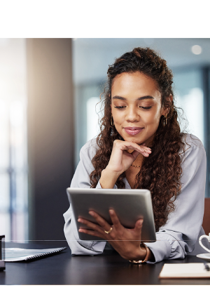 Woman sitting at a desk with a coffee cup and notebook, smiling while looking at a tablet.